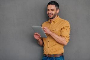 Examining his brand new tab. Handsome mature man holding digital tablet and looking at it with smile while standing against grey background photo