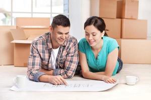Examining a house plan. Cheerful young couple lying on the floor and looking at the blueprint while cardboard boxes laying on the background photo