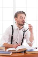 Waiting for inspiration. Thoughtful young man in shirt and suspenders holding glasses and looking away while sitting at his working place photo