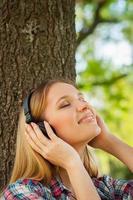 disfrutando de la música en el parque. vista lateral de una hermosa joven con auriculares escuchando música y sonriendo mientras se apoya en el árbol en un parque foto