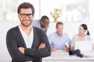 Team leader. Handsome young man in glasses keeping arms crossed and smiling while three people working on background photo