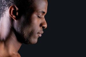 calmado y seguro. vista lateral de un joven africano que mantiene los ojos cerrados mientras se enfrenta a un fondo gris foto