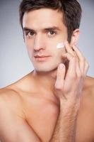 Skin care. Handsome young shirtless man applying cream at his face and looking at camera while standing isolated on grey background photo