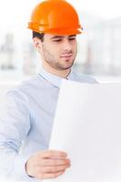 Examining a blueprint. Confident young man in shirt and tie examining blueprint while standing against a window at office photo