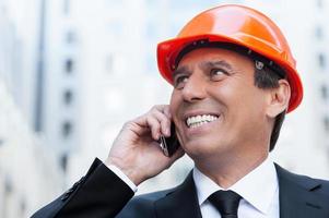 Happy contractor. Cheerful mature man in formalwear and hardhat talking on the mobile phone and smiling while standing outdoors photo