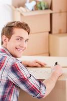 Marking a carton box. Top view of cheerful young man marking a cardboard box and looking over shoulder while more boxes laying in the background photo