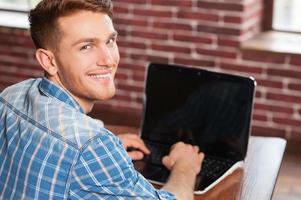 Young and creative. Rear view of handsome young man working on laptop and looking over shoulder while sitting at his working place photo