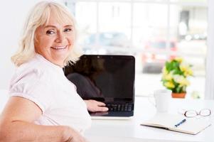 Feeling young and full of energy. Rear view of cheerful senior woman looking over shoulder and smiling while working on laptop photo