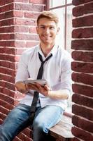 Handsome with digital tablet. Cheerful young man in shirt and tie working on digital tablet and looking at camera while sitting at the window sill photo