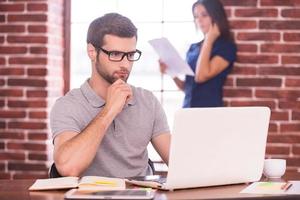 Doing creative work. Confident young man working on laptop while woman standing in the background and talking on the mobile phone photo