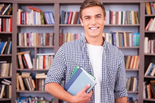 Student in library. Handsome young man holding books and smiling while standing in library photo