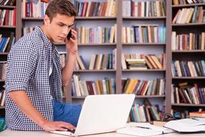 plena concentración en el estudio. un joven confiado que trabaja en una laptop y habla por teléfono móvil mientras se apoya en el escritorio y frente a la estantería foto