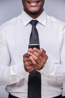 Businessman with smart phone. Cropped image of happy young African man in shirt and tie holding smart phone and smiling while standing against grey background photo