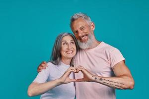 Beautiful senior couple looking at camera and showing heart with hands while standing together against blue background photo