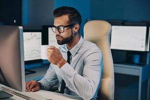Confident young man drinking coffee and working on computer while staying late in the office photo