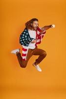Confident young man in casual clothing making a face and punching while hovering covered in American flag against yellow background photo