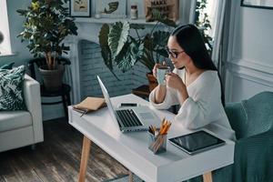 Planning another step. Top view of beautiful young woman working using computer and smiling while sitting in home office photo