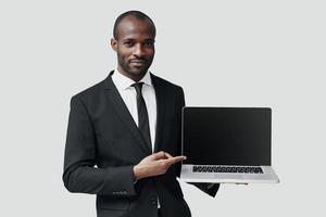 confiado joven africano en ropa formal señalando espacio de copia en la computadora portátil y sonriendo mientras está de pie contra el fondo gris foto