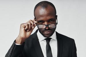 Confident young African man in formalwear looking at camera and adjusting eyewear while standing against grey background photo