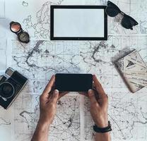 New adventure. Close up top view of man using smart phone with sunglasses, photo camera, compass and passport lying on map around