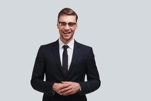 Confident business expert. Handsome young man in full suit keeping hands clasped and looking at camera with smile while standing against grey background photo