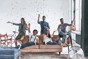 Private party.  Group of happy young people in casual wear drinking champagne while spending time at home with confetti flying everywhere photo