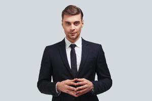 Young perfectionist. Handsome young man in formal wear keeping hands clasped and looking at camera with smile while standing against grey background photo