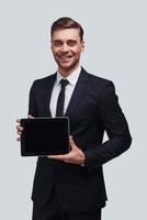 Using modern technologies. Good looking young man in full suit holding digital tablet and looking at camera while standing against grey background photo