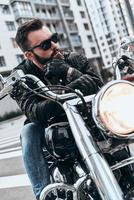 luciendo genial apuesto joven con chaqueta de cuero y gafas de sol montando en moto mientras pasa tiempo al aire libre foto