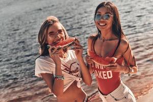 relajación de verano. vista superior de mujeres jóvenes atractivas sonriendo y comiendo sandía mientras están de pie cerca del río foto