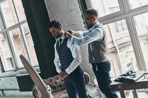 Paying attention to every detail. Young fashionable designer helping his client to get dressed while standing in his workshop photo