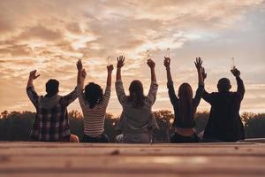 Friendship that lasts forever. Rear view of young people in casual wear toasting with a beer bottles while sitting on the pier photo
