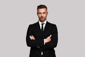 Business expert. Handsome young man in full suit keeping arms crossed and looking at camera while standing against grey background photo