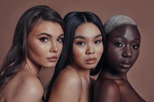 Calm and beautiful. Three attractive young women looking at camera while standing against brown background photo