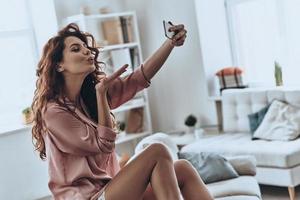 Sharing photos. Attractive young women blowing a kiss while taking selfie at home photo