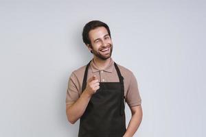 Handsome young man in apron looking at camera and pointing you while standing against gray background photo