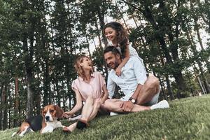 Enjoying every moment together. Happy young family of three with dog smiling while sitting on grass in park photo