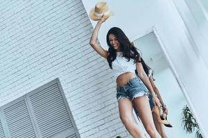 She is all about style. Attractive young woman holding a sun hat beyond her head and smiling while standing at home photo