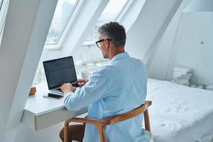 Rear view of confident mature man working on laptop while sitting at the office desk photo