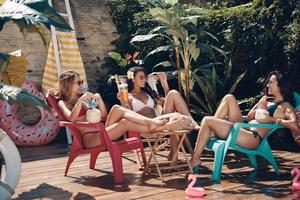 mujeres jóvenes atractivas en traje de baño bebiendo cócteles y sonriendo mientras se relaja en la piscina al aire libre foto