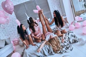 Crazy ideas. Playful young women trying to put on bunny ears on their girlfriends foot and smiling while sitting on the bed photo