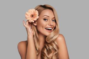 hermosa mujer joven ajustando la flor en su cabello y sonriendo mientras está de pie contra el fondo gris foto