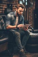 Checking appointment list. Handsome young bearded man working on digital tablet while sitting in comfortable chair at barbershop photo