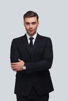 Confident in his style. Handsome young man in full suit keeping arms crossed and looking at camera while standing against grey background photo