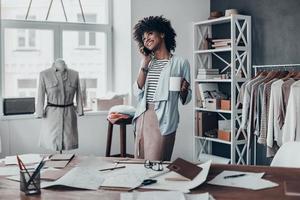 hablando con un cliente feliz. atractiva joven africana hablando por teléfono inteligente y sonriendo mientras está de pie en su taller foto