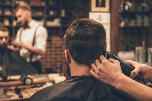 Nape trim. Rear view of young bearded man getting haircut by hairdresser with electric razor while sitting in chair at barbershop photo