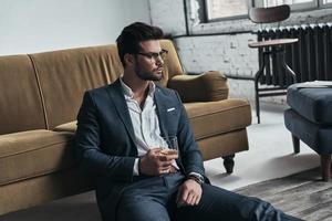 Enjoying favorite drink. Handsome young man in full suit holding a drinking glass and looking away while sitting on the floor at home photo