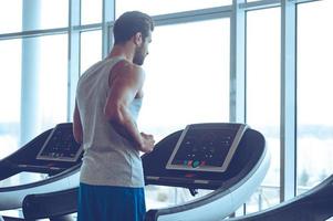 Body power. Young man in sportswear running on treadmill in front of window at gym photo