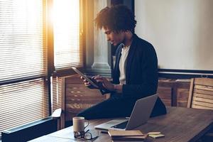 Managing his timetable. Side view of young handsome African man using his digital tablet while sitting on table at his working place photo