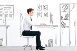 New working day. Side view of handsome young man working with drawn computer while sitting at his illustrated working place photo